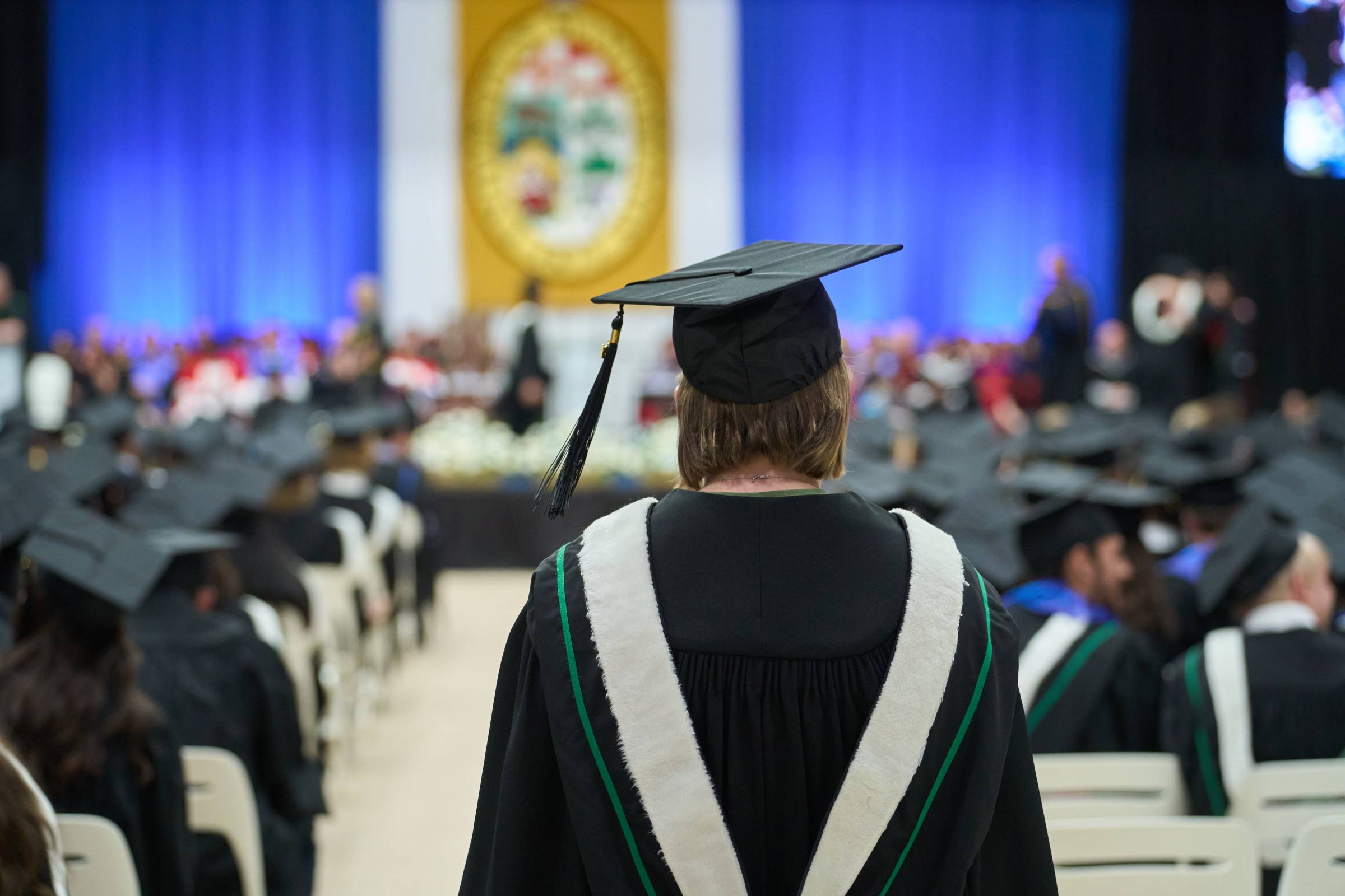 A grad in cap and gown walks down the centre aisle at Convocation. A banner with the University Crest hangs on the wall in the distance.