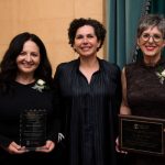 Three people pose for a photo. Two of them are holding award plaques.