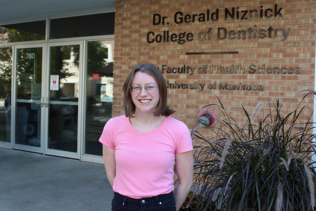 Portrait of Kayla Austin. She is standing outside of a building. On the wall behind her reads "Dr. Gerald Niznick College of Dentistry, Rady Faculty of Health Sciences, University of Manitoba. "