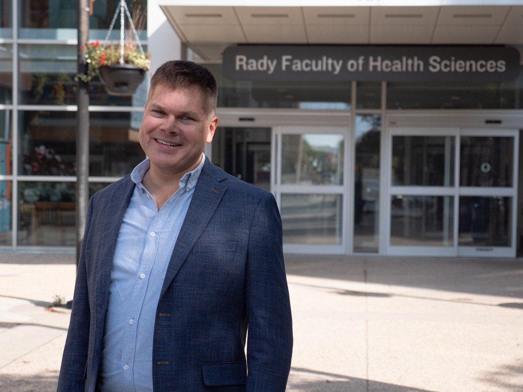 Barret Monchka smiles at the camera while standing outside the Brodie Centre. In the background, the entrance to the Rady Faculty of Health Sciences is visible.