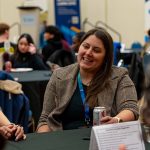 Female employer sitting at a round table with students, wearing a lanyard, smiling and chatting.