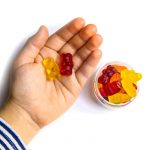 A bowl of gummy bears sits beside a child's hand holding two gummy bears.