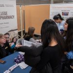 Two people sit behind a table at a College of Pharmacy booth. Three students stand in front, chatting with them.