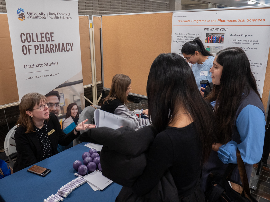 Two people sit behind a table at a College of Pharmacy booth. Three students stand in front, chatting with them.