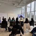 A large group of people sitting in a room watching the presenter on a TV who is presenting remotely.