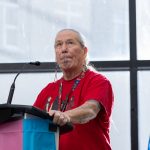 Elder Charlotte Nolin speaks at a podium, which has a trans flag on front.