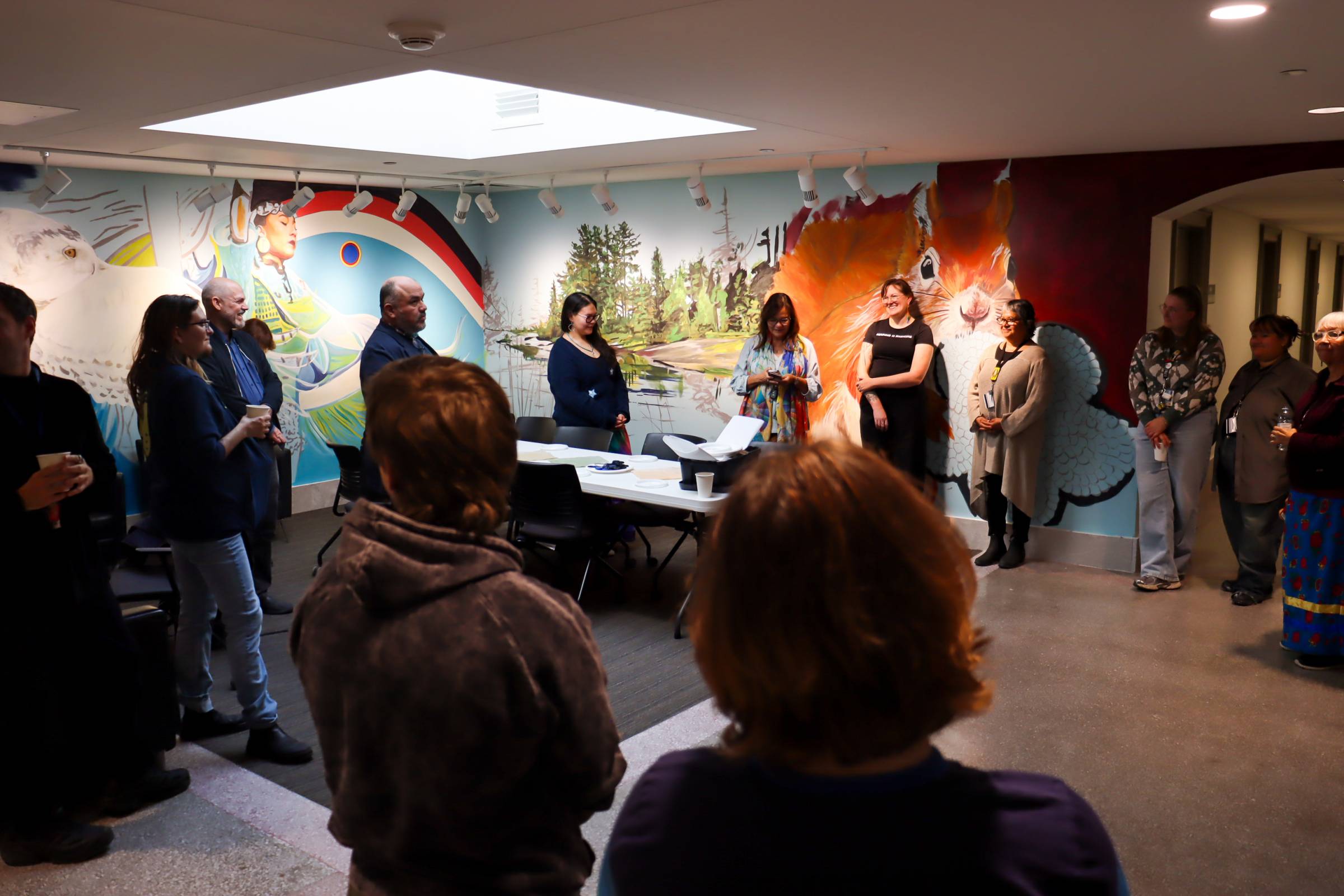 Students and community members gather in the Indigenous Student Space for opening remarks, surrounded by a mural.
