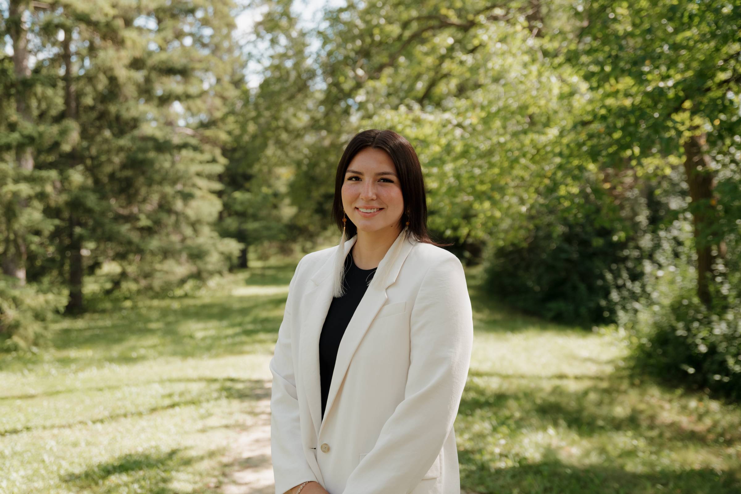 Tréchelle Bunn in a white blazer standing in an open green space surrounded by trees, sunshine and blue sky.