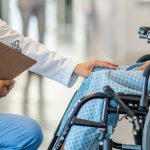 A patient sits in a wheelchair and is helped by a doctor who approaches patient in an empathetic way.